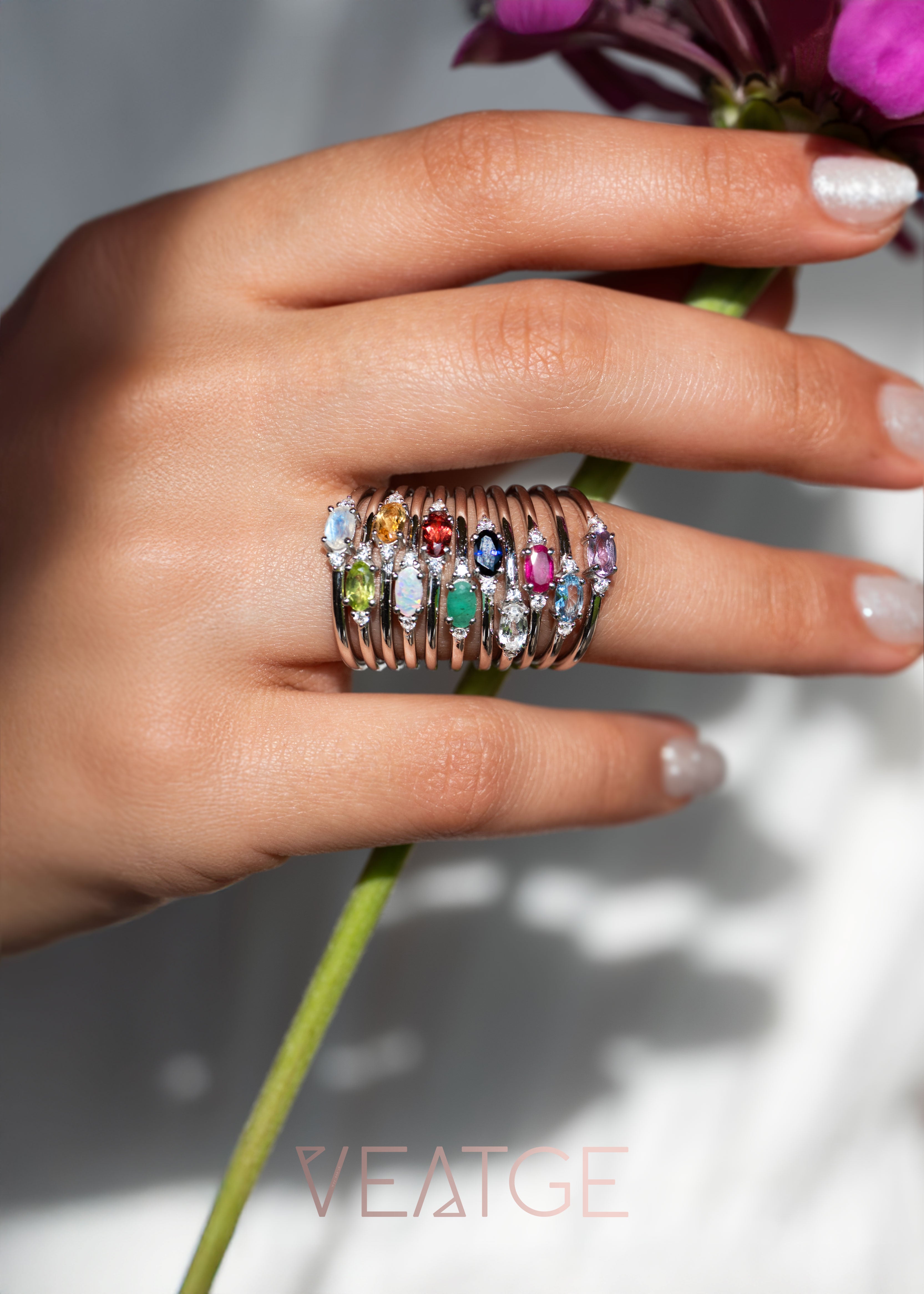 Hand wearing a stack of colorful gemstone rings holding flowers with 'VEATGE' branding.