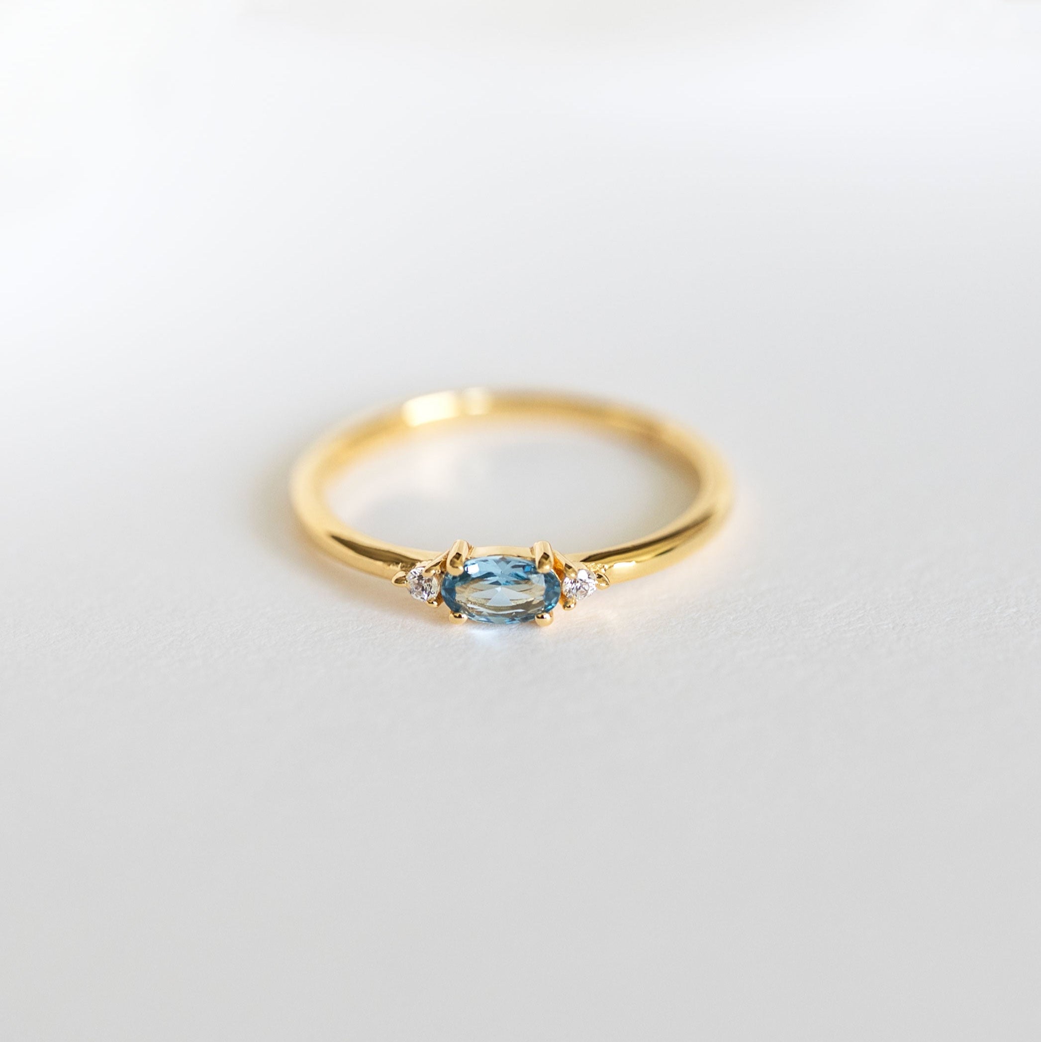 A gold vermeil dainty stacking ring with a marquise cut aquamarine center stone and two smaller clear stones on the sides, branded 'VEATGE'.