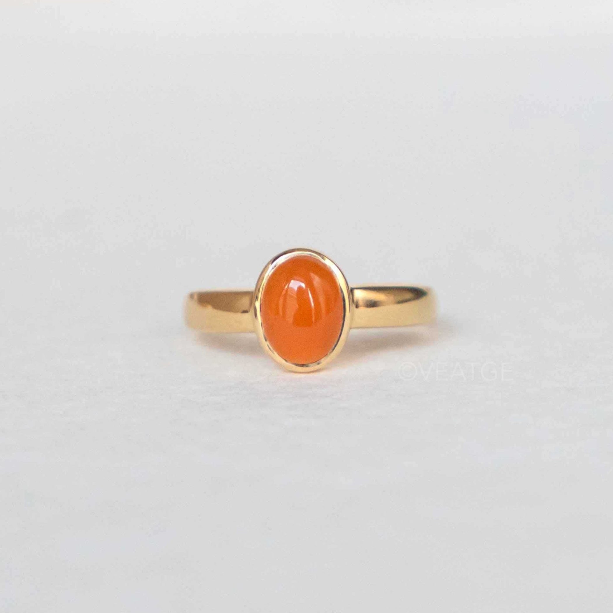 A carnelian ring with a clementine hue, set in a bezel setting on a 925 sterling silver base with a gold plating, presented on a white background with the brand name 'Veáge' visible in the corner.