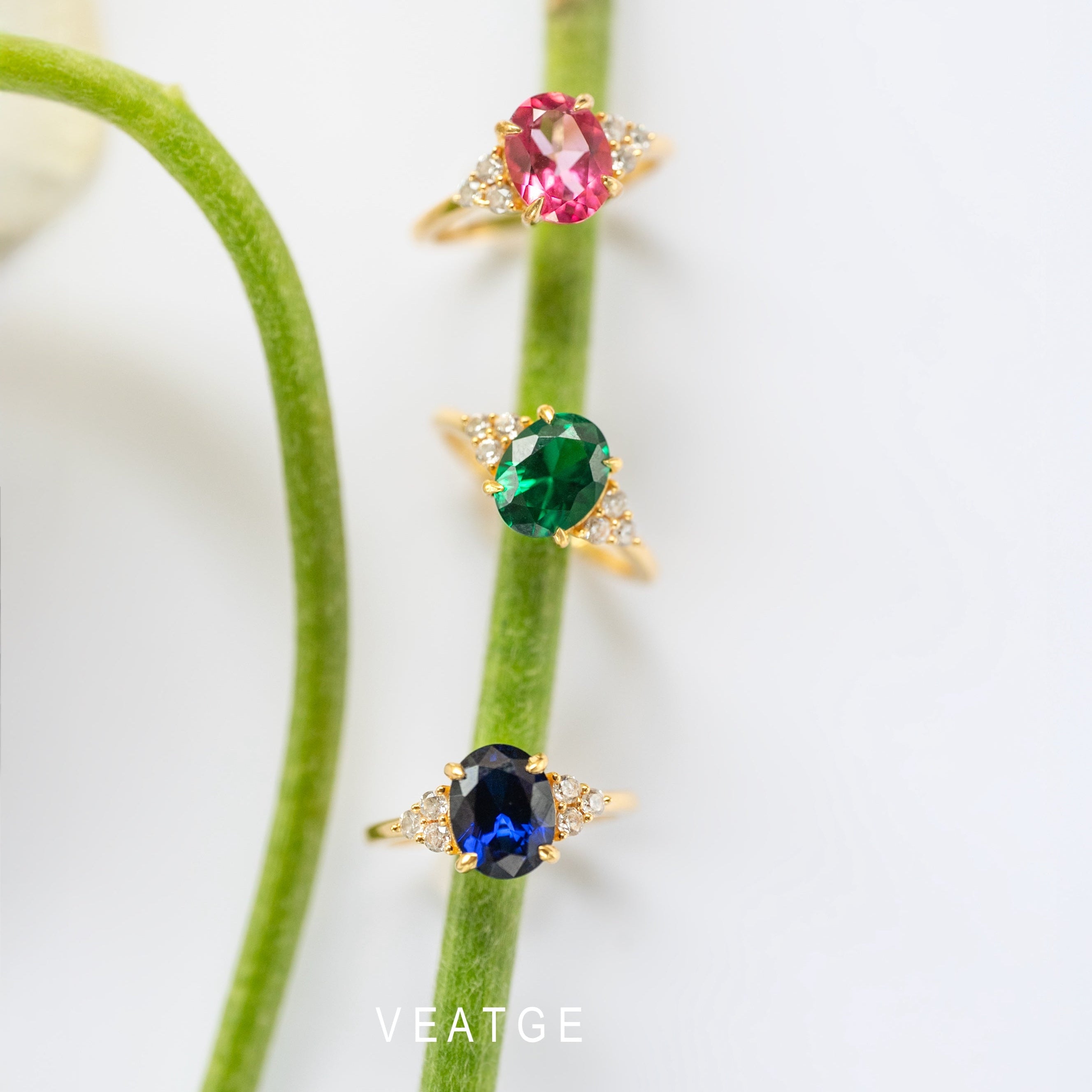 Three gold rings with different colored gemstones on a green stem against a white background.