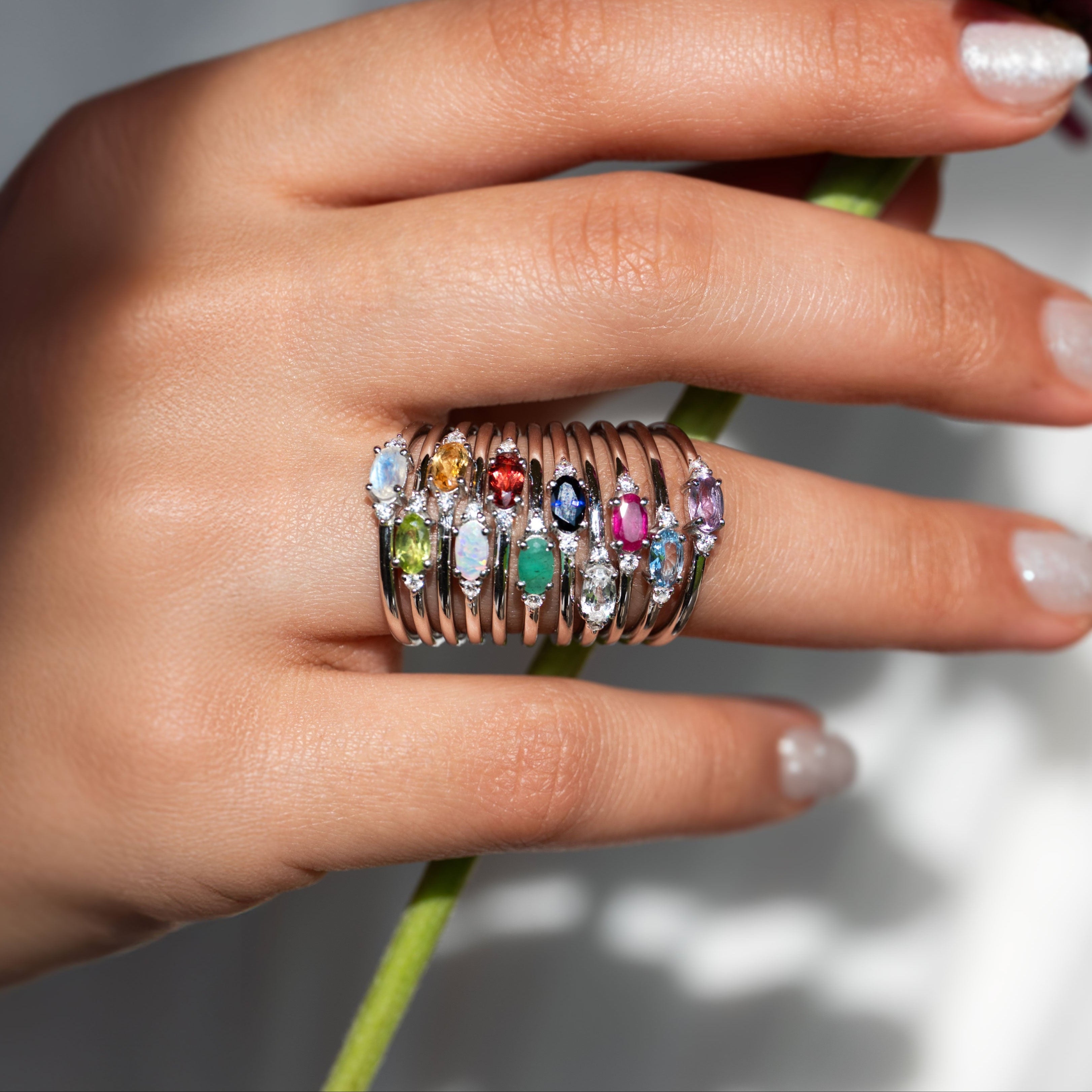 Hand wearing a stack of colorful gemstone rings holding flowers with 'VEATGE' branding.